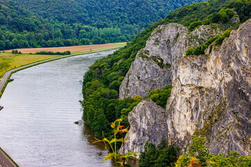 Meuse River and rock formation known as Freyr Rocks, among meadow, hills with abundant green leafy trees, irregular rock wall famous place for climbing, cloudy summer day in Wallonia, Belgium © Emile