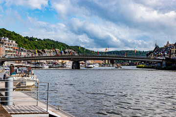 Meuse river with boats anchored and sailing, vehicular bridge with waving flags in Dinant city, cityscape with buildings on background, sunny summer day in Namur province, Wallonia, Belgium