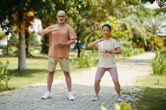Multiracial man and woman practicing Tai Chi in a serene park setting. Both engaging in synchronized movements among lush greenery, enjoying fitness in nature