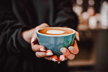 A close-up of two hands holding a blue ceramic cup with a latte, featuring intricate latte art. The warm beverage and cozy atmosphere create a comforting and inviting scene, perfect for coffee lovers.