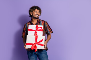 Fototapeta premium Photo of cheerful man with wavy hair dressed print shirt holding gift boxes look at promo empty space isolated on purple color background