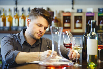 Male sommelier tasting red and white wine, making notes at bar counter