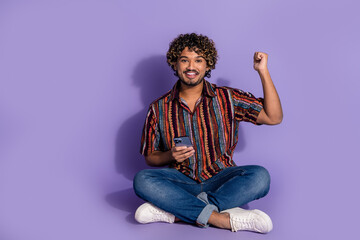 Full size photo of satisfied ecstatic man dressed print shirt jeans sit hold smartphone win lottery isolated on purple color background