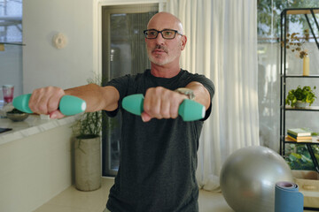 Man standing in living room exercising with dumbbells and wearing glasses and dark shirt, a fitness ball and white curtains visible in background