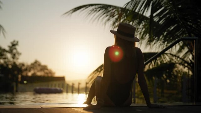 Young lady in hat splashing water of pool by legs sitting on edge, back view. Female tourist admiring sunset in summer evening, rest in luxury hotel in holidays and vacation, silhouette, rear view