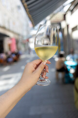 A close-up of a hand holding a glass of white wine in an outdoor cafe, with a blurred background featuring people and outdoor seating in a lively atmosphere.