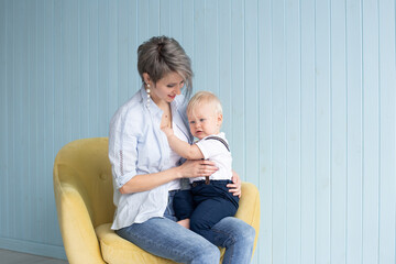 Young mother with baby son, while sitting on armchair at home near a blue wooden wall, happy family