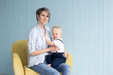 Mother with baby son, while sitting on armchair at home near a blue wooden wall, happy family