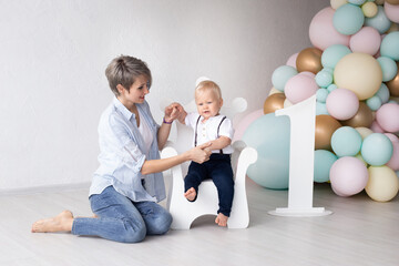 Mother with her baby boy at home on his first birthday sits on a throne-shaped chair, with the number one next to him in the background of colorful balloons.
