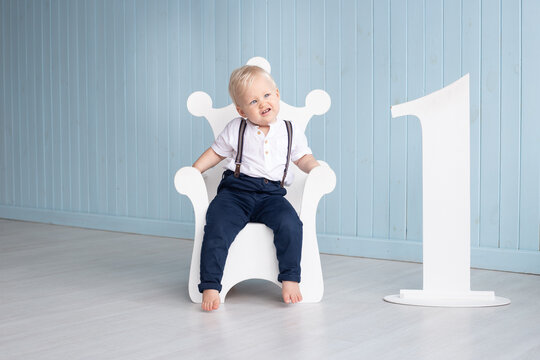 Little boy on his first birthday sits on a throne-shaped chair, with the number one next to him, dressed in a suit with trousers on pull-ups and a shirt near blue wall at home