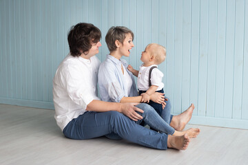 Happy family, mother, father and their son baby boy hugging at home while sitting on floor near blue wall in casual clothes