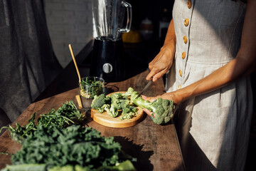 Female hands with knife, broccoli, herbs and blender. Young woman is cutting cabbage for green smoothie or salad in kitchen at home.