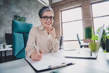 Photo of smiling positive mature lady accountant formalwear writing papers indoors workplace...