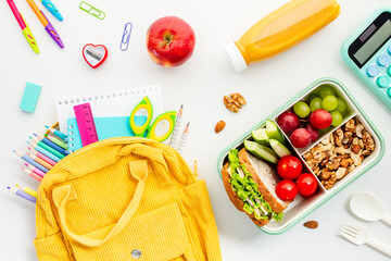 Flat lay yellow backpack, school supplies, a healthy lunchbox, and fresh fruits on a white...