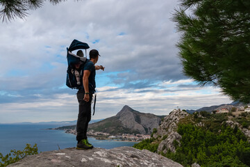 Hiker man with baby carrier backpack on rock formation with aerial view of coastal town Omis,...