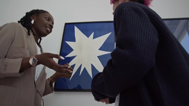 Low angle shot of young Black woman discussing modern artwork with Asian friend while standing together in exhibition hall of art gallery