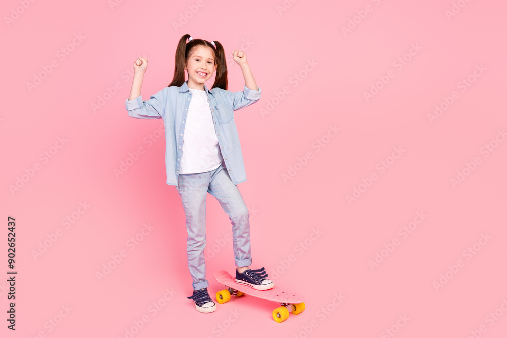 Poster Full size photo of positive small girl dressed denim shirt pants stand on skateboard clenching fists isolated on pink color background