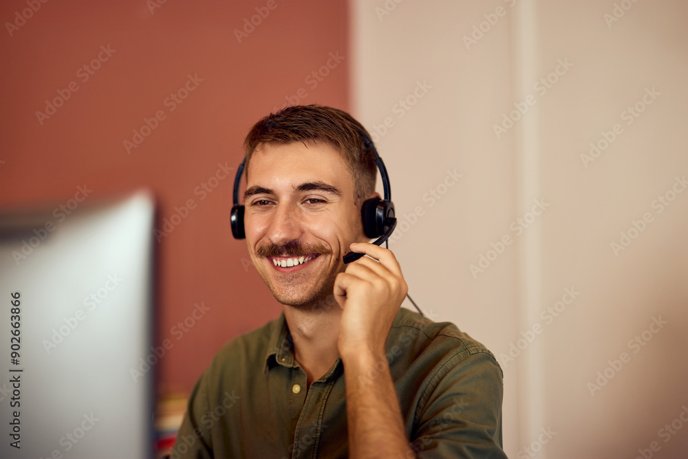 Wall mural A smiling man using headphones with a microphone, working over the computer, having an online meeting.