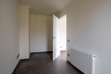 An empty room with clean walls and tiled flooring, featuring an open door that lets light in and a white radiator beneath the window, creating a minimalistic space.