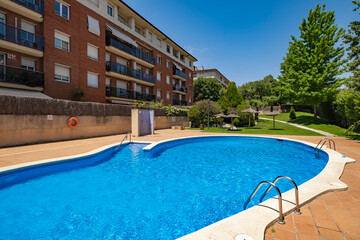 An outdoor swimming pool with clear blue water, surrounded by apartment buildings and green garden areas, under a bright blue sky on a sunny day.