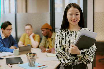 Medium portrait of cheerful young Asian female specialist holding documents and digital tablet standing in office and smiling at camera