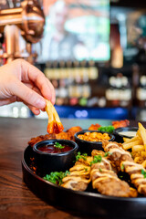 Close-up of a hand dipping a french fry into sauce with a plate of assorted snacks in the background at a bar. Perfect for illustrating casual dining, food enjoyment, and social gatherings.