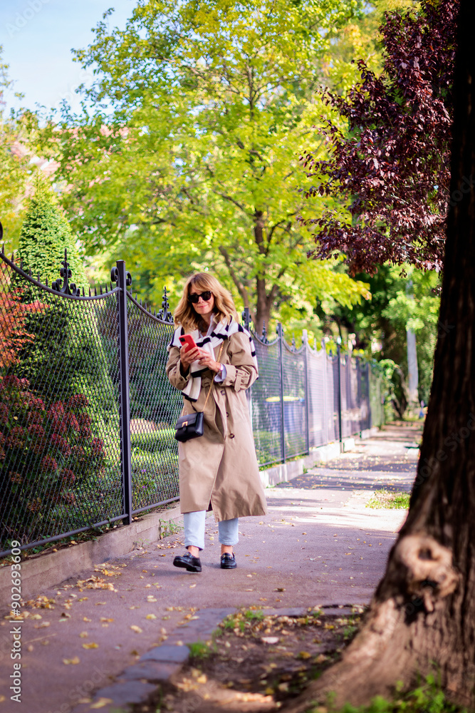 Wall mural Full length of a smiling blond haired woman using smarpthone and walking down the street