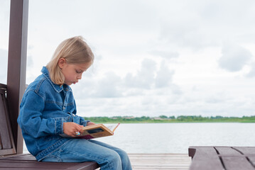 Blonde girl reads a book while sitting in a gazebo on the shore of a lake.