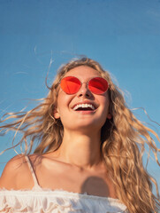 A woman with long blonde hair, wearing red-tinted sunglasses, smiling brightly against a clear blue sky