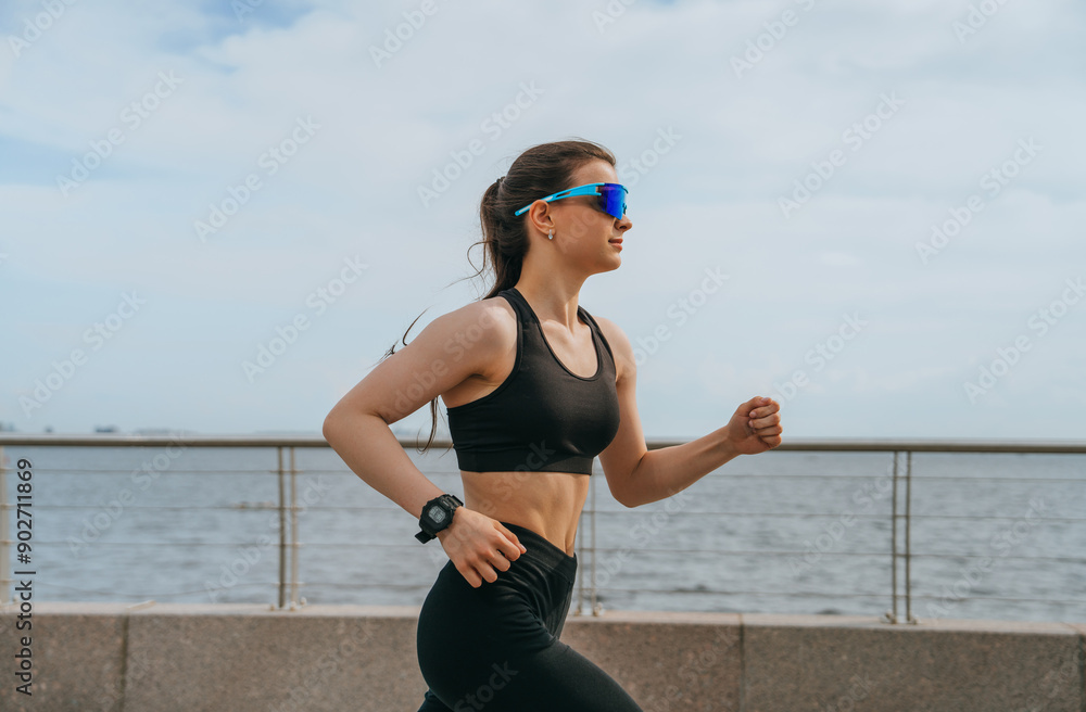 Wall mural young woman in black sportswear jogging by the water, wearing sunglasses and a watch. the focus is o