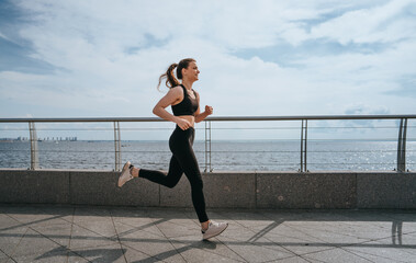 young woman jogging along a waterfront path, dressed in black sportswear. The sky is partly cloudy,...