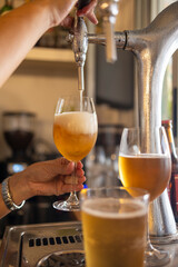 A close-up shot shows a hand expertly pouring beer with a thick foam head into a wine glass from a tap, set against a background of a bar counter with another filled glass nearby.