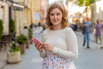 Smiling happy redhead young woman using smartphone typing text email messages browsing internet app, finishing work, looking at camera outdoors. Girl walking in urban city street. Town lifestyles.