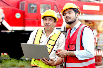 Two workers with safety vest and helmet hold laptop computer for discussion at construction site. Senior engineer and labor inspecting building site. Mature elderly and young architect work together.