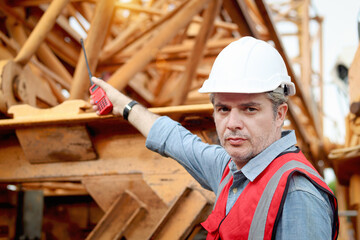 Portrait of senior worker engineer foreman with safety vest and helmet holds walkies talkie and pointing at construction site. Mature elderly architect working at ground level of building workplace.