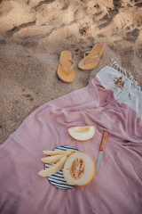 A pink blanket lies on the sand by the sea. On it is a cut melon on a striped plate and a knife...