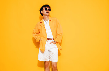 Young attractive man with short curly hair hairstyle. Smiling handsome male in casual stylish shirt and shorts clothes posing in studio, isolated on yellow. Cheerful and happy model, sunglasses