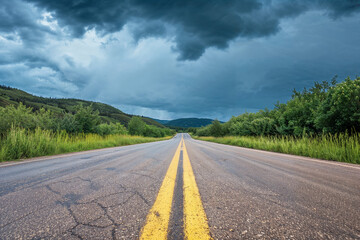 a deserted road with a yellow centerline stretching into the distance, flanked by greenery under a dark, ominous sky filled with heavy storm clouds