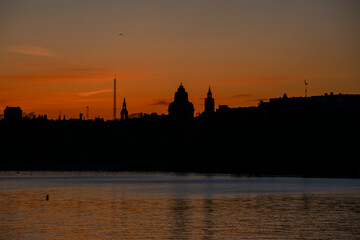 Naklejka premium Panoramic view of Stockholm City hall at sunset with orange sky and calm water reflection. Scenic skyline of the Swedish capital at dusk. 