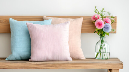 A bench with three pillows and a vase of flowers. The pillows are pink, blue, and white. The bench is wooden and has a natural feel. The scene is peaceful and calming
