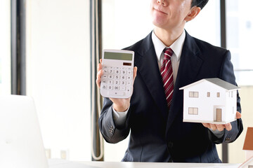 住宅模型と電卓を持ち悩むスーツ姿のアジア人男性のビジネスパーソン