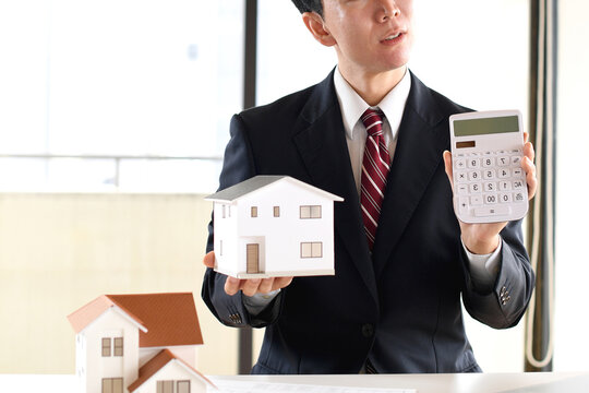 住宅模型と電卓を持ち悩むスーツ姿のアジア人男性のビジネスパーソン