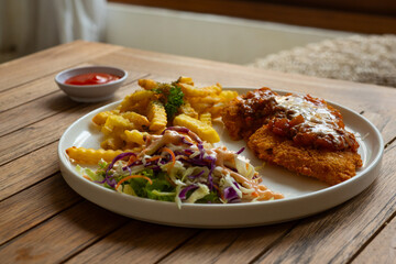 A plate of chicken parmesan or chicken parmigiana, served alongside french fries and salad. On a rustic wooden table, in cafe environment
