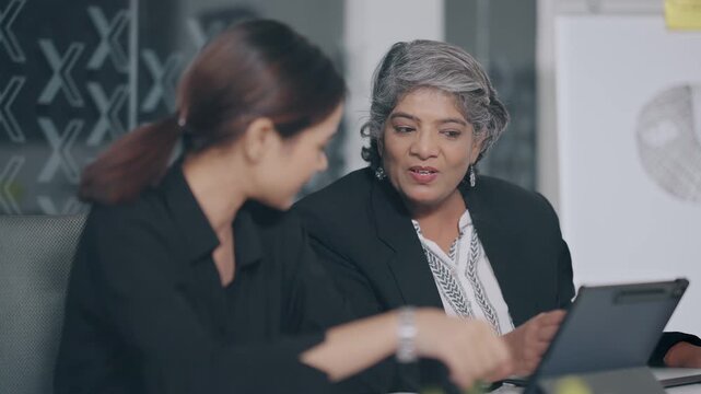 Indian older aged boss women use tablet touch pad look screen talk new deal plan share ideas with teen girl indoor job office young adult expert female old elder lady staff do work day task work place