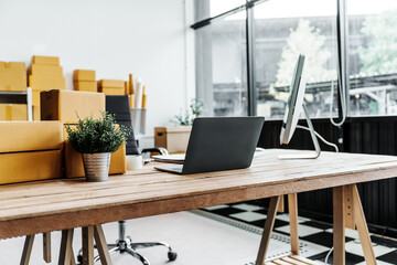 A wooden desk in an SME office, with a laptop, desktop computer, and a parcel cardboard box. The workspace is organized for efficient management, logistics, and order processing.