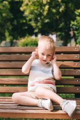 A boy with fair hair makes faces while sitting on a bench in a summer park.