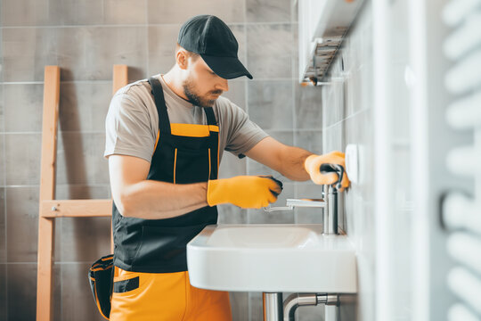 Young handsome plumber in uniform fixing pipes under sink: bathroom home service, professional handyman maintenance job