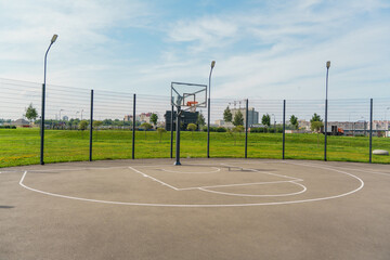 Outdoor basketball court in a sunny park, surrounded by greenery and a fence, under a clear blue sky.