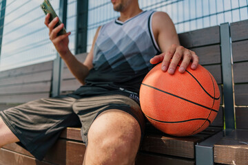 A basketball player seated on a bench, holding a basketball and checking his phone during a break.