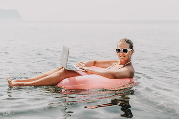 A woman is sitting on a pink inflatable raft in the ocean, holding a laptop. She is smiling and she is enjoying her time. Concept of relaxation and leisure.